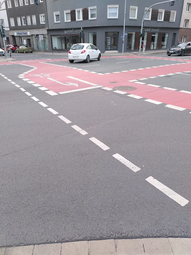 Eine Kreuzung zweier mehrspuriger Straßen. Der Beobachter steht auf dem Bürgersteig an einer Ecke der Kreuzung. Auf der Kreuzung sind in Rot Radwege eingezeichnet. Für jede Richtung gibt es eine Einbuchtung für indirektes Linksabbiegen.