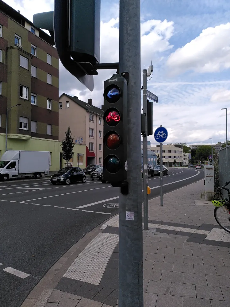 Eine Fahrradampel in Nachaufnahme. Die Ampel ist rot. Über dem roten Ampellist ist ein weiteres Ampelelement mit einem blass weiß leuchtenden Pfeil.
