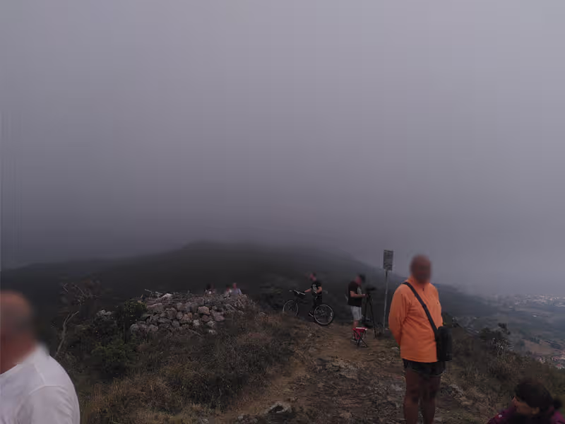 Im Vordergrund mehrere Menschen, die auf einem spärlich bewachsenen Untergrund stehen. Es ist neblig, im Hintergrund kann man noch eine Stadt und einen anderen Hügel erkennen. Die Spitze des Hügels ist schon im Nebel verschwunden.