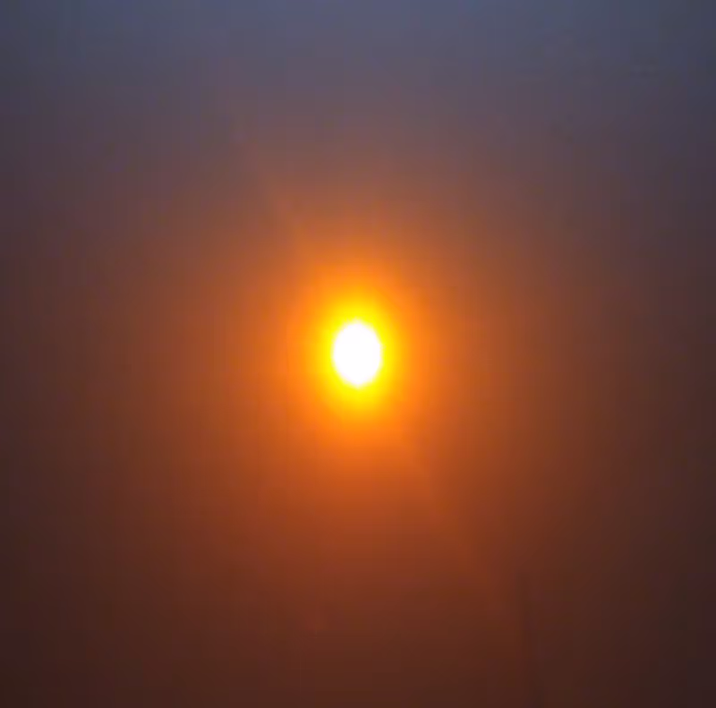 Eine weiße Ellipse, die nach außen hin zunächst gelb, dann orange wird bis sie schließlich in den dunklen Hintergrund übergeht. Quer durch den weiße Fleck geht eine Linsenreflektion.