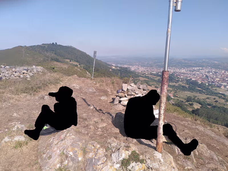 Zwei ausgeschwärzte Personen sitzen auf felsigem, mit vertrocknetem Gras bewachsenen Untergrund. Es ist sonnig, der Himmel ist blau. Im Hintergrund sind rechts grün bewachsene Hügel, links eine Stadt im Tal und eine grüne, leicht ausgetrocknete Landschaft.