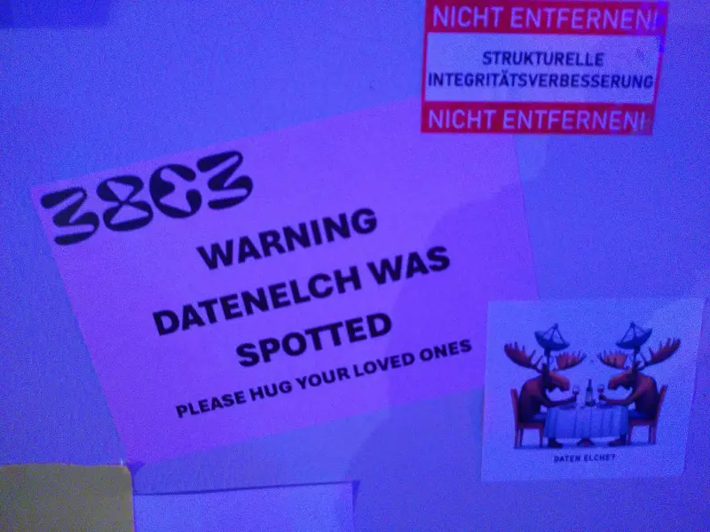 Ein Aushang. Dort steht: „WARNING DATENELCH WAS SPOTTED PLEASE HUG YOUR LOVED ONES“. Daneben ein Aufkleber mit zwei Datenelchen, die an einem Tisch sitzen und ein Glas Wein trinken.