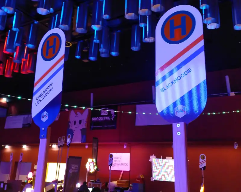 Zwei Schilder, die wie Bushaltestellenschilder aussehen. Sie sind beschriftet mit „Chaosdorf Düsseldorf“ und „Blackhoodie“. Im Hintergrund sind weitere solcher Schilder.
