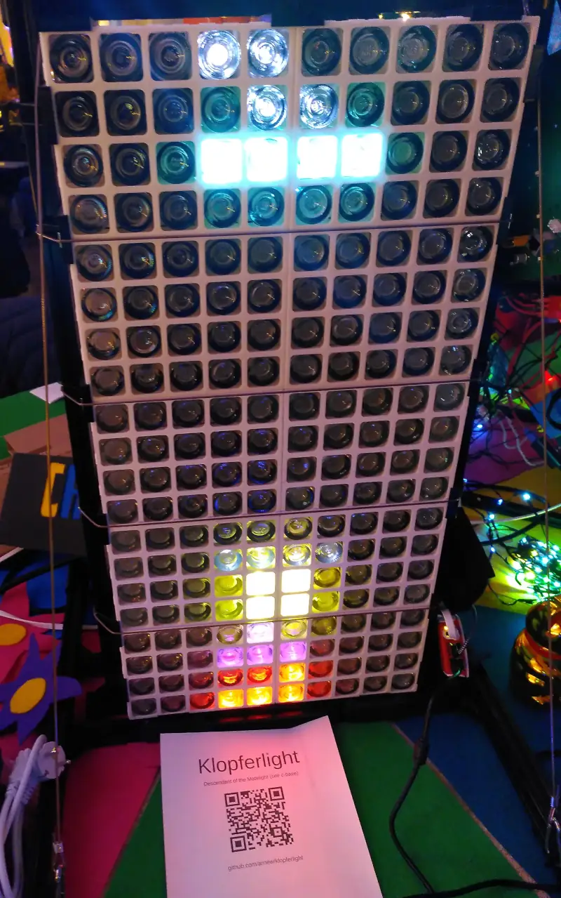 Eine Matrix aus Fläschchen, mit den Flaschenböden nach vorne. Die meisten Flaschen sind dunkel, ein paar sind in verschiedenen Farben beleuchtet. Darunter ein Ausdruck mit der Aufscrift „Klopferlight“ und einem QR-Code.