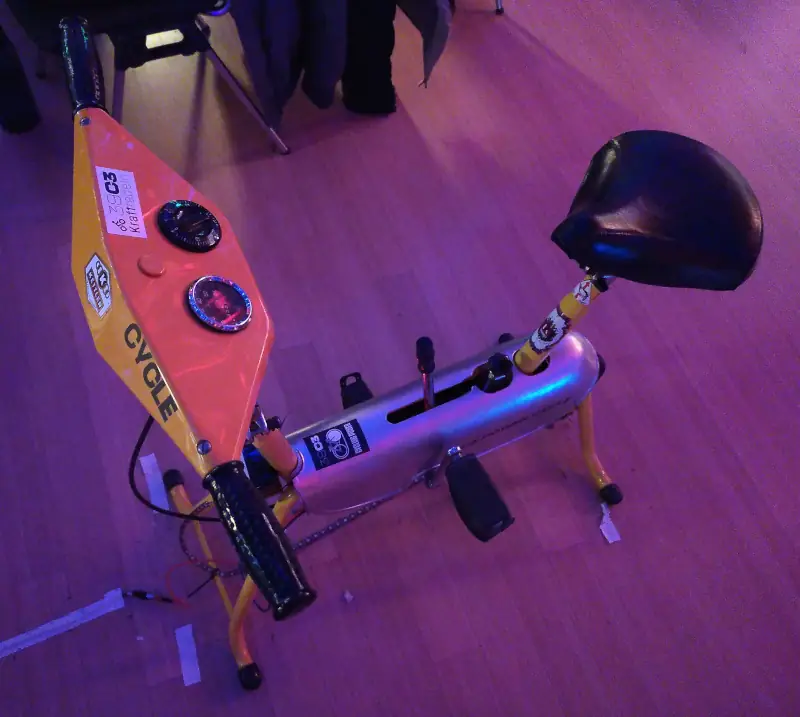 Ein Fahrradergometer. Auf dem gelb lackierten „Lenker“ ist ein Aufkleber mit der Aufschrift „CYCLE“