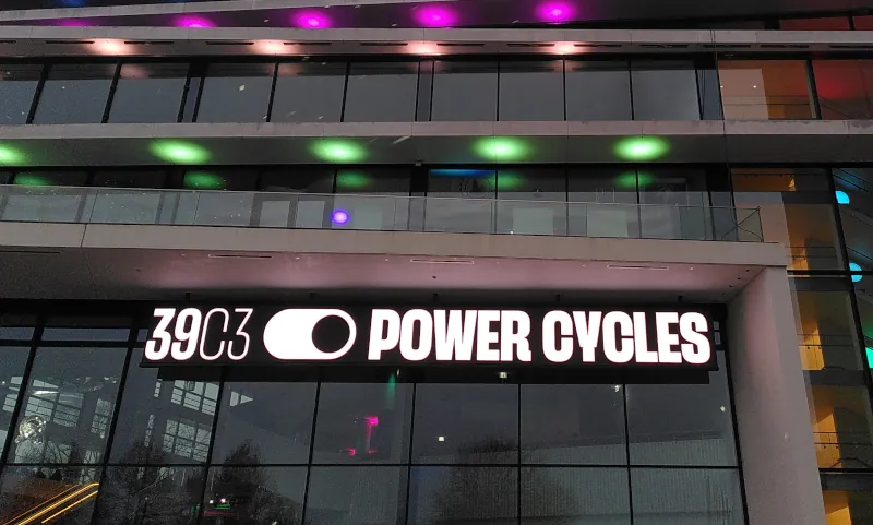Eine Fassade mit vielen Glasscheiben und bunten Lichtern. An der Fassade ist eine große Leuchttafel mit der Aufschrift „39C3 POWER CYCLES“