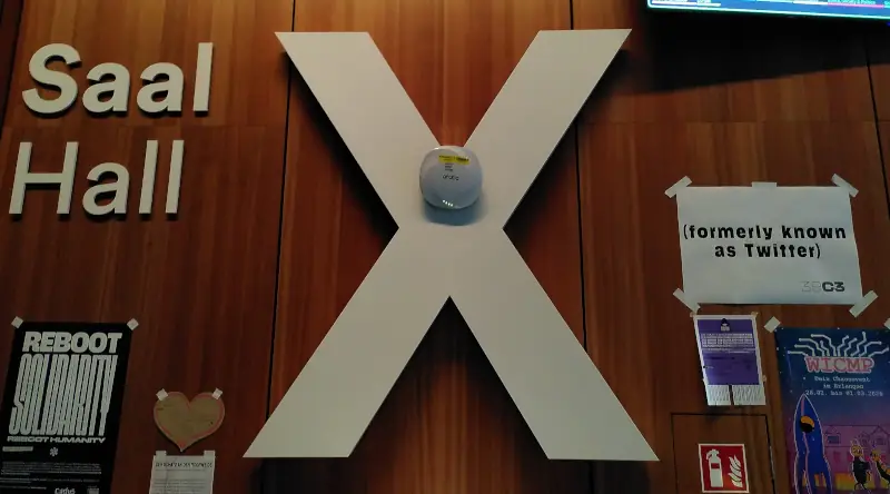 Eine Holzvertäfelung. Dort steht in großen weißen Buchstaben „Saal X“. Daneben hängt ein DIN A4-Zettel mit der Aufschrift „(formerly known as Twitter)“