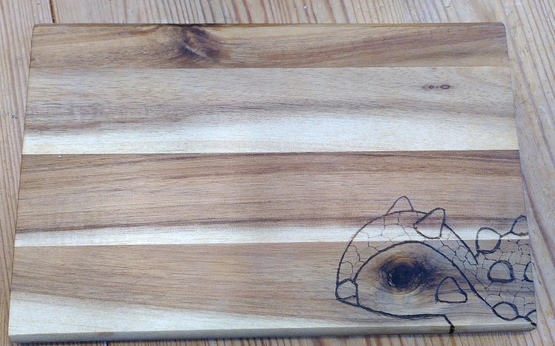 Ein Holzbrettchen. Unten rechts sind Kopf und Schultern eines Ankylosaurus eingraviert (Brandmalerei / Pyrographie). Ein Astansatz im Holz dient als Auge des Sauriers.
