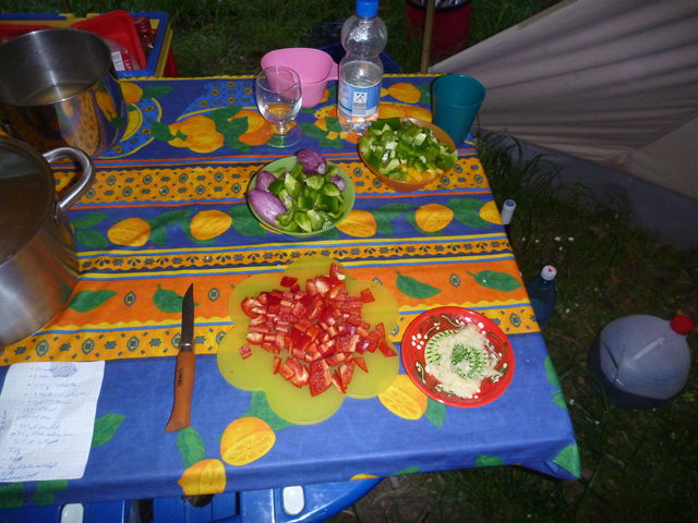 geschnittene Paprika in mehreren Schälchen und auf einem Brettchen, geriebener Knoblauch auf einer Keramikreibe. Alles steht auf einem Campingtisch.