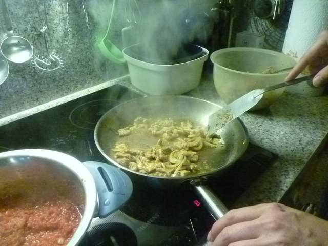 Eine Pfanne, in der das Fleisch gebraten wird. Von der Pfanne steigt Dampf auf. Im Bild sind zwei Hände zu sehen, eine Hält die Pfanne, die andere einen Pfannenwender.