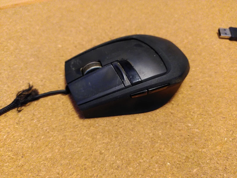 Eine schwarze Computermaus auf einer Korkunterlage. Die Maus hat mehr Tasten als üblich. Sie ist etwas dreckig und abgenutzt.