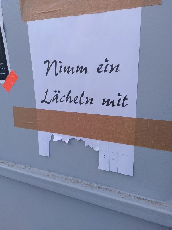 An eine Wand geklebt ist ein DIN A4-Zettel mit der Aufschrift „Nimm ein Lächeln mit“. Darunter, mit Einschnitten, Ascii-Smilies zum Abreißen. Einige davon sind schon abgerissen.