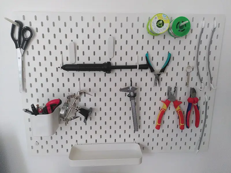 An einer Wand hängt eine weiße Platte mit im Raster angeordneten senkrechten Schlitzen. In einigen dieser Schlitze hängen Behälter oder Aufhängungen. Daran hängen u.a. eine Greifzange, ein Seitenschneider, Rollen mit Lötzinn, eine Scheere und Stifte. Einige Halterungen sind noch frei.