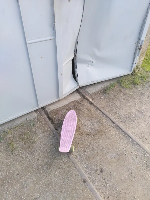 Ein kleines pinkes Skateboard steht verlassen vor einer Blechwand. Die Blechwand ist stark verbogen. Die Spitze des Skateboards zeigt auf die Blechwand.