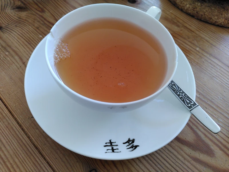 Eine weiße Porzellantasse gefüllt mit Schwarztee. Sie steht auf einer weißen Untertasse, auf der zwei chinesische Schriftzeichen zu sehen sind. Neben der Tasse, auf der Untertasse, ist der Griff eines Teelöffels zu sehen.