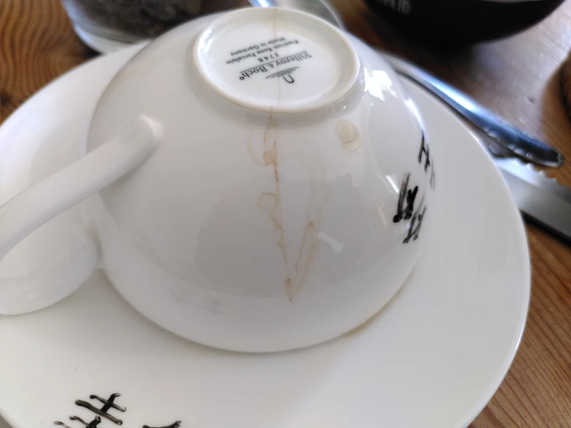 Eine weiße Teetasse, die umgekehrt auf einer Untertasse liegt. Durch die Tasse zieht sich ein Riss. Vom Riss aus gehen eingetrocknete Teetropfen. Auf Tasse und Untertasse sind chinesische Schriftzeichen.