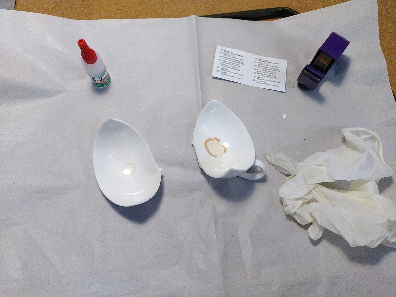Eine graue Papierunterlage. Auf ihr liegen mehrere Gegenstände: eine in zwei Hälften zerbrochene weiße Teetasse, ein Kleberfläschchen, eingeschweißte Reinigungstücher, ein Tesafilmabroller und ein Paar Einweghandschuhe