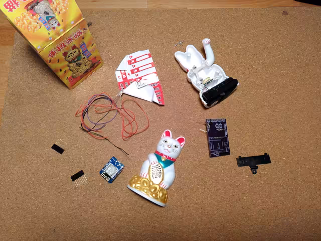 Auf einer Korkoberfläche liegen verschiedene Teile: Kabel, zwei LEDs, ein PCB (auf dem schon ein paar Teile aufgelötet sind), ein Microcontroller und zwei Hälften einer Plastikwinkekatze.